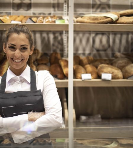 portrait-of-bakery-seller-with-arms-crossed-standing-in-front-of-shelf-full-of-bred-bagels-and-pastry-min